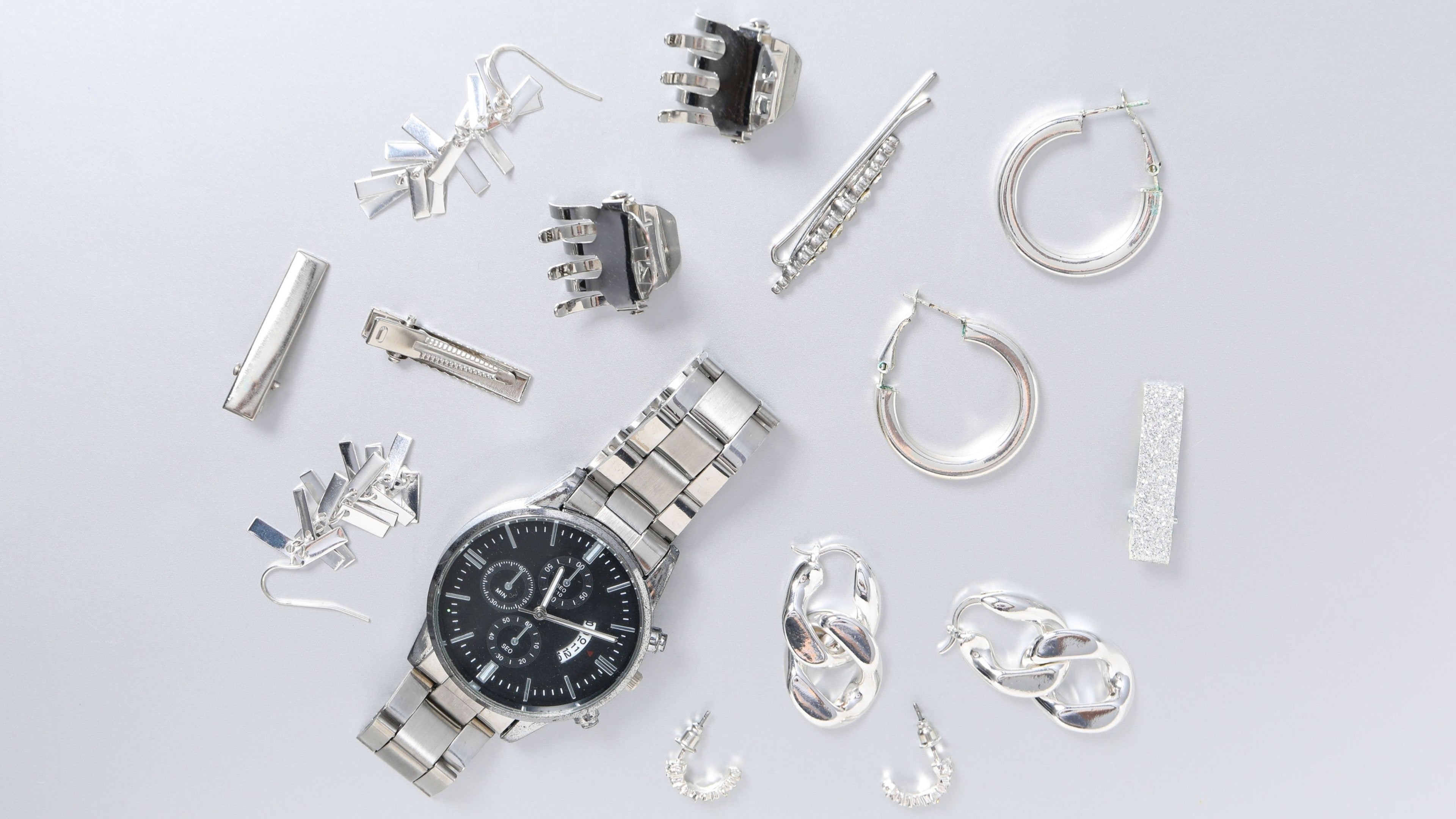 Flat lay of a silver wristwatch surrounded by various silver earrings, clips, and jewelry pieces on a light surface.