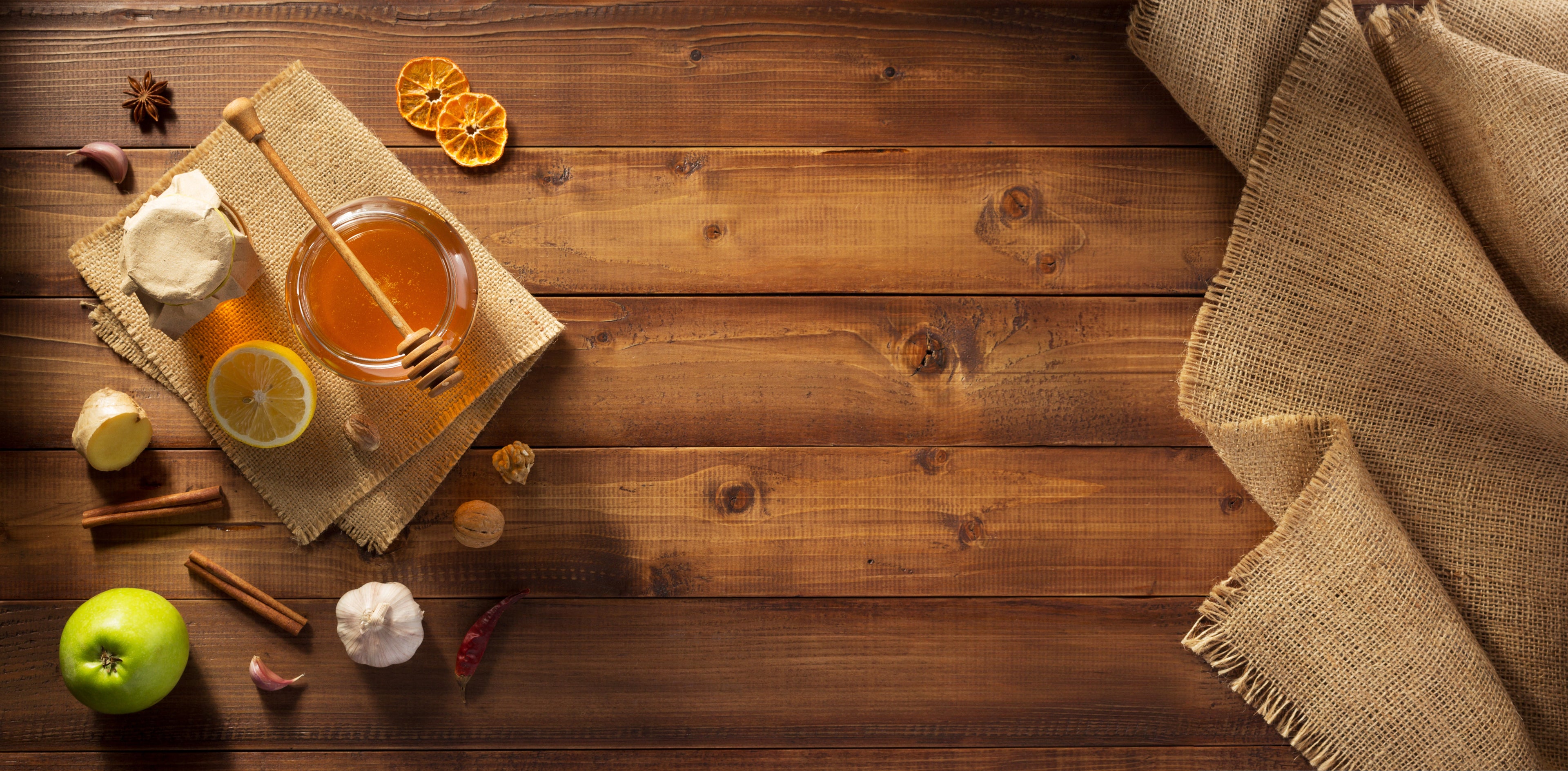 Assortment of natural food ingredients including honey, lemon, ginger, garlic, spices, and a green apple on a wooden table.