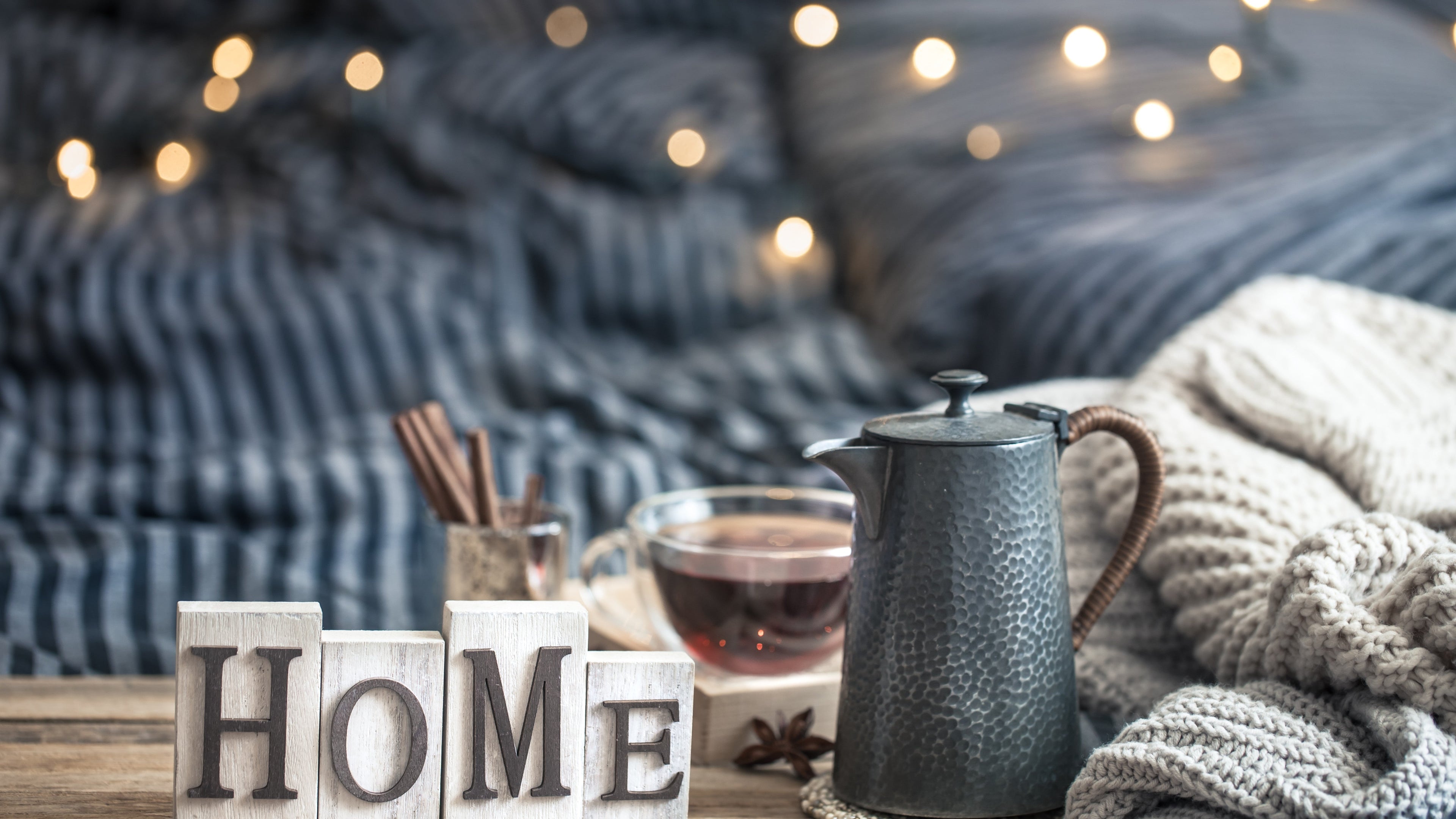 Cozy home setting with a metal teapot, cup of tea, knitted blanket, cinnamon sticks, and wooden blocks spelling “HOME” against soft string lights.