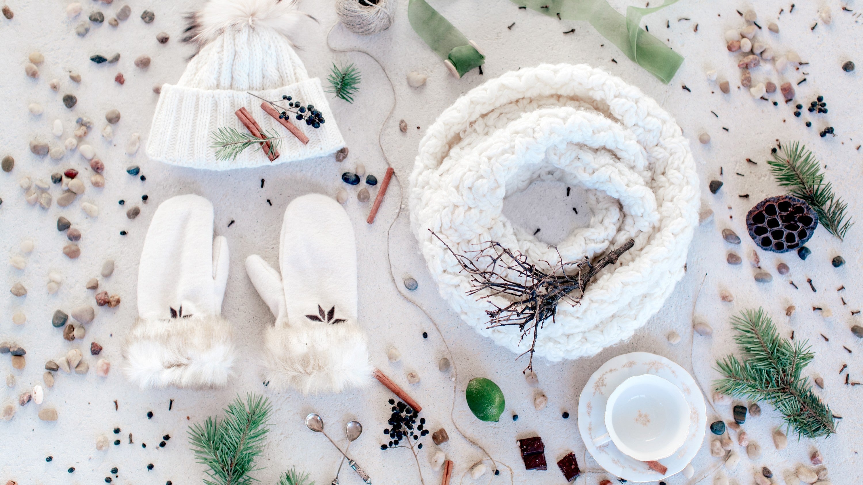 Flat lay of winter accessories including a white knit hat, white mittens, and a chunky knit scarf surrounded by pine branches, cinnamon sticks, berries, stones, and winter-themed decorations.